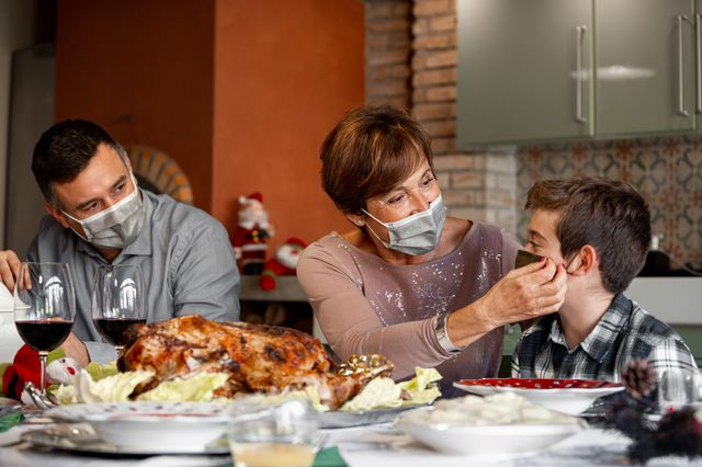 A stock photo shows a man and a woman wearing masks as the woman adjusts a boy's mask at a dinner table with a turkey.